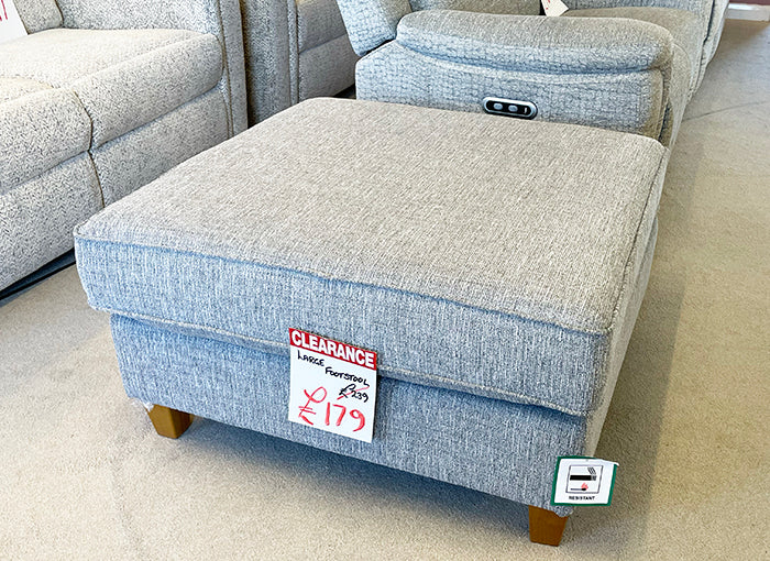 Large grey upholstered footstool with a clearance sign on a carpeted floor