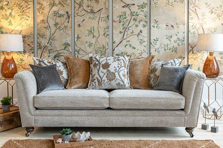 A beige Alstons sofa from the Inca collection with cushions against a floral patterned wallpaper.