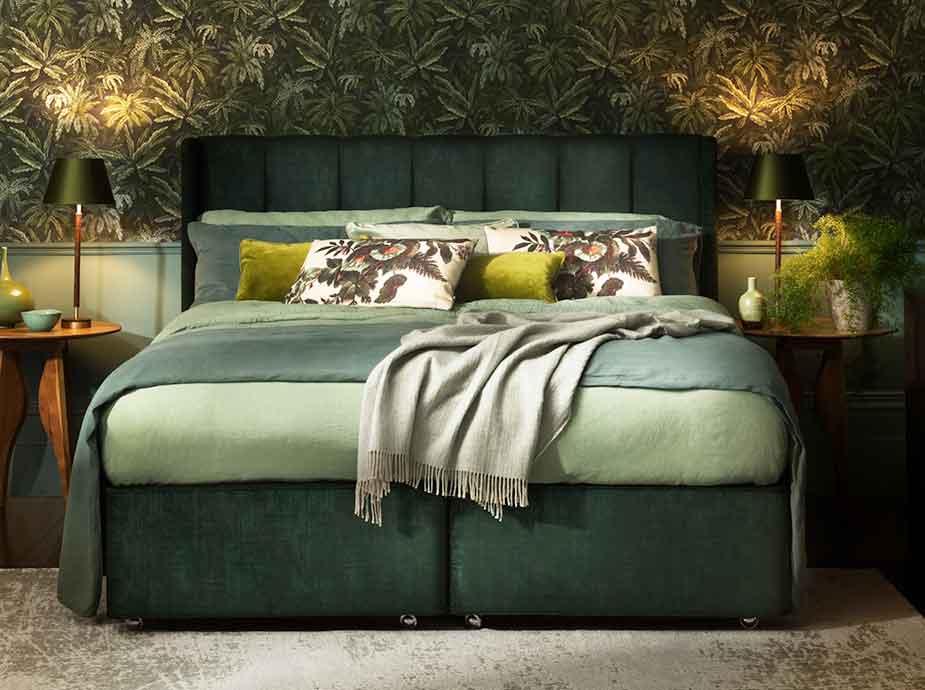 Green upholstered bed with decorative pillows against a leafy wallpapered wall.